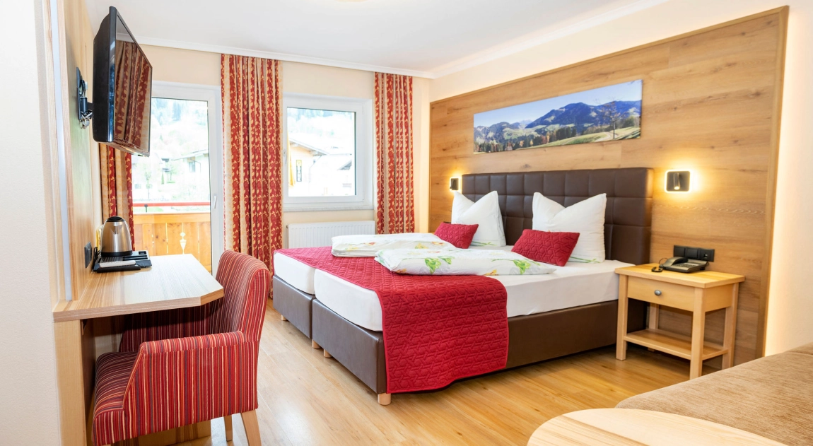 Helles Hotelzimmer mit Doppelbett, Schreibtisch, TV, roten Vorhängen und großem Fenster mit Blick nach draußen.
